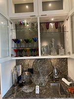 Open cabinet displaying multiple shelves of coloured stemmed glasses, clear crystal decanter and glasses, silver cocktail shaker, ice bucket, and mixing glasses on countertop.