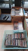 Photo showing two boxes and a plastic container filled with multiple rows of CDs seen from above, packed vertically with titles legible from spines.