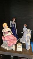 Four ceramic figurines displayed together on a shelf, showing variety of dresses and poses.