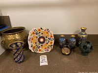 Photo showing Asian decorative lot items including a brass planter, twisted blue glass vase, multi-color floral ceramic plate, two small cloisonné vases, black planter, and green Chinese puzzle ball.