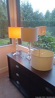 Two clear acrylic bodied table lamps with white drum shades placed on dark wood dresser near window with greenery outside.