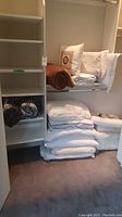 Closet view showing assorted white pillows stacked on the floor and a brown throw blanket rolled on a shelf.