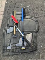 Four black rubber car mats arranged flat on the ground, three snow brushes placed horizontally on top of the mats on a paved surface with visible cracks.
