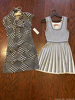 Two dresses laid flat on wooden floor. Left dress is black and white geometric pattern with wide collar. Right dress is white sleeveless knit with black vertical stripes and flared skirt.