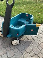 Image of teal Step2 children's wagon with door open, showing two-seat interior, black pulling handle, and four white wheels on paved ground with grass background.