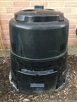 Black plastic compost bin with screw-on lid and front door, standing outdoors against a brick wall. Visible ventilation slots and some wear on surface.