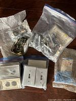 Assorted electrical hardware including wire nuts, metallic switch components, plastic switch plates, duplex outlets, and switches in plastic bags on wood floor.