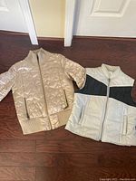 Beige quilted jacket and beige/black faux leather vest laid out flat on floor, showing front details including zippers and collars.