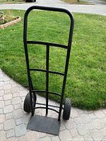 Front view of black metal hand truck dolly with large pneumatic tires on a paved patio with grass in the background.