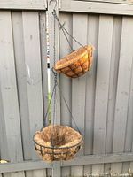 Set of two metal hanging planters with coconut coir liners hanging against an outdoor fence. Shows the design and materials of the baskets and liners.