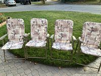 Four collapsible patio chairs with built-in floral cushions on metal frames, placed in an outdoor setting on paving stones with grass and driveway in background.