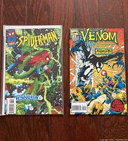 Photo showing Spider-Man Media Blizzard Part 3 and Venom Symbiote Unleashed comics side by side on a wooden floor, both in protective sleeves.