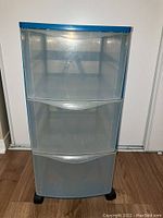 Front view of blue frame storage tower showing three translucent drawers with handles and black wheels at base.