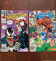 Two Amazing Spider-Man comic books shown flat on wood surface, one with Venom holding a skull with Spidey mask and the other with Spider-Man surrounded by other characters including Sandman.