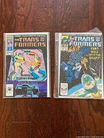Photo of two vintage Transformers comic books issue #5 and another issue with 'Fort Max Gets the Shaft!' cover art, sleeved with backer-board on a wooden floor.