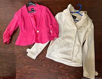 Lot contains two dressy women's jackets laid flat on wood floor, one pink blazer with single gold button and ruched sleeves, one cream-colored double-breasted peacoat jacket.