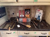Barbecue tools including long forks, tongs, oil brush and bags of smoking chips and wood chunks arranged on kitchen counter.