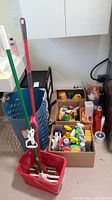 Overall photo showing blue rolling cart, red mop bucket with mop, and cardboard boxes containing cleaning bottles and sprays