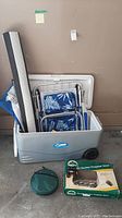 Wheeled cooler filled with foldable camp chair, rolled camping mat, boxed propane stove, and green umbrella case on concrete floor against wall.