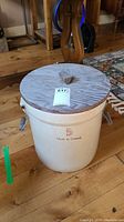 Full view of ceramic crock with wooden lid and metal side handles on wood floor.