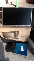 Acer B246HL monitor standing on its box with two other items in front: a black Verbatim USB 1TB external drive and a blue Brickell 6710U scale.