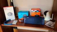Front view of all lot items on wooden shelf: August lock box, blue and orange watch bands, Echo Dot box, Sony speaker, Fire TV Cube box, D-Link camera and manual.