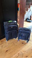 Two black soft-sided rolling suitcases standing upright on hardwood floor near dark furniture and stairs, showing overall size comparison