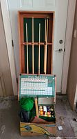 Four wooden pool cues on a wall-mounted rack with green felt, box of pool balls and accessories visible below.