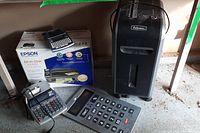Wide shot showing Fellows 99Ci shredder, Epson CX8400 printer box, Brother label maker on top, Staples large calculator, and Canon MP11DX calculator side by side on the floor.
