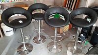 Four black faux leather upholstered bar chairs on chrome bases shown together. Adjustable height and swivel features visible.
