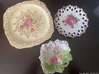 Three decorative plates laid flat showing floral designs and detailing including gold accents and pierced edges.