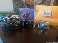 Photo showing three cameras with their original boxes: Nikon One Touch 100, Canon PowerShot SX120 IS, Canon PowerShot A560.