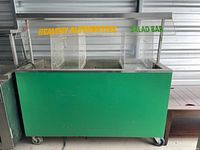 Green cold buffet salad bar with labeled glass sneeze guard and multiple food compartments visible on top.