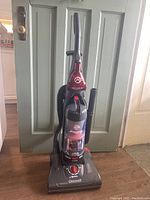 Full view of the Dirt Devil Jaguar Pet upright vacuum cleaner showing red and black coloring, hose, and clear dirt canister.