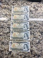 Five Canadian one dollar bills laid out side by side on a granite surface, showing front faces with Queen Elizabeth II portrait and clear serial numbers.