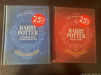 Front cover of two hardcover Harry Potter books with 25% off stickers, one blue title 'Harry Potter Character Compendium', one red title 'Harry Potter Bestiary'