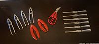 Top-down view of all utensils in the set arranged in a line, showing four silver-toned shell crackers, two red plastic shell crackers, one pair of scissors, and six seafood forks.