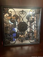 Square mirror featuring an ornate metal design overlay frame with central medallion and floral scroll motifs, placed on a wood surface.