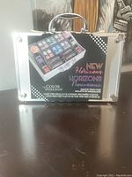 Front view of closed silver hard makeup train case labeled The Color Workshop New Horizons with product description and picture.