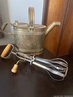 Image showing brass watering can with handle and spout diagonally positioned next to vintage hand mixer with crank and wooden handles.