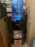 Full view of the water cooler unit with blue water jug on top, placed against wall in corner.