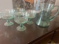 Photo showing the full set of one green glass water pitcher and four matching green glass pedestal glasses with embossed patterns.