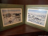 Two framed winter scenery pictures resting on a wooden surface, showing snowy village scenes with people ice skating and snowy backgrounds.