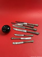 Seven knives with ornate sterling silver handles and stainless steel blades displayed on red background, including four large knives and three butter knives. Green apple used for size reference.