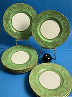 Group of 11 green and gold gilt dinner plates displayed stacked and on easels alongside a 3-inch reference apple.