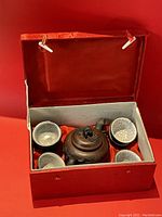 Open presentation box displaying teapot and six matching cups inside red cloth lining.