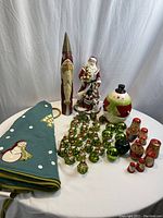 Wide view of the entire Christmas lot on a white cloth table showing the frog ornaments, additional glass ornaments, wooden Santas, ceramic snowman cookie jar, ceramic Santa, carved wooden Santa, and green tree skirt.