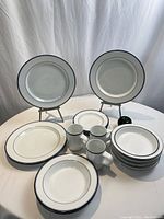 Photo showing the full set on a white tablecloth with two dinner plates on stands, bread plates, salad plates stacked, soup bowls stacked, and three coffee mugs in front. A green apple is included for size reference.