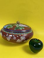 Front angle showing floral design on lid and body with brass knob on lid, next to a green apple for size reference.
