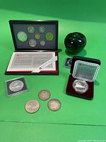 Royal Canadian Mint coin set displayed in black and red case with informational inserts, plus three loose antique silver coins and two large encased silver coins on green background with green glass apple for scale.